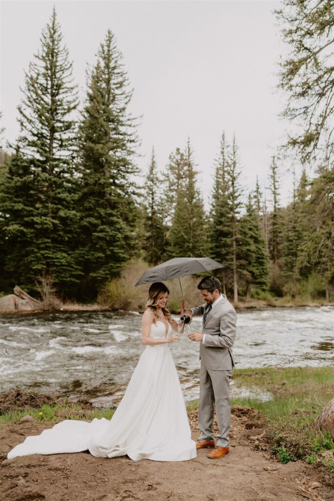 couple pouring glasses of champagne by a creek while holding an umbrella as it rains
