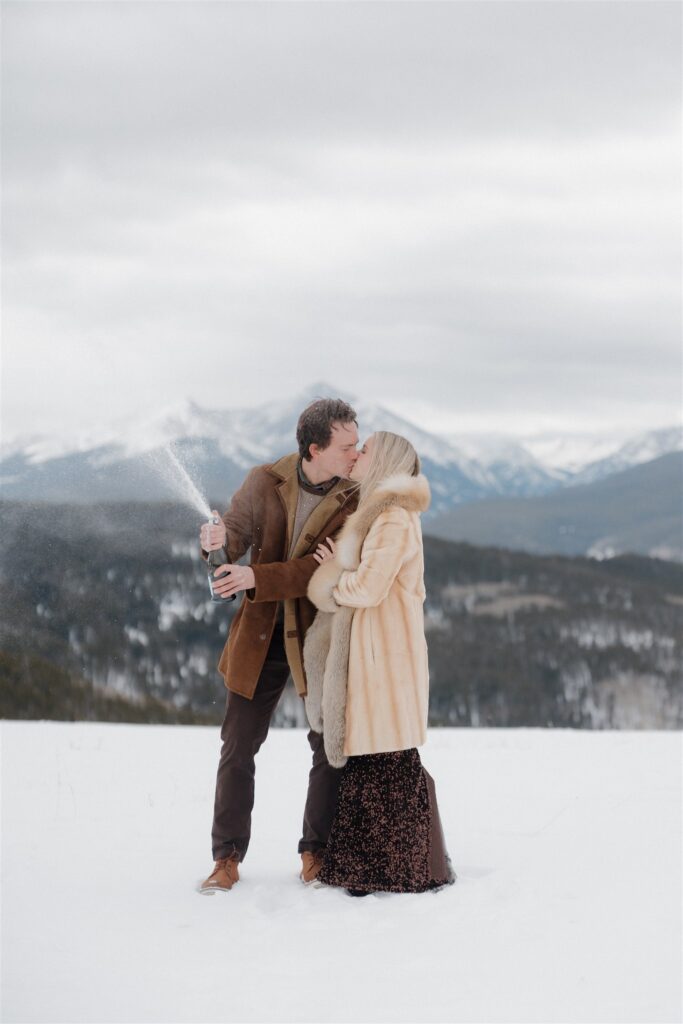 Couple pops champagne during their snowy engagement photos